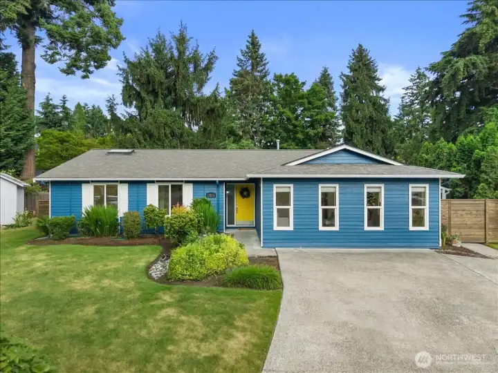 Front Exterior – Single-level home with blue exterior siding, white trim, and a bright front entry door. Landscaped front yard features mature shrubs, lawn space, and a wide driveway.