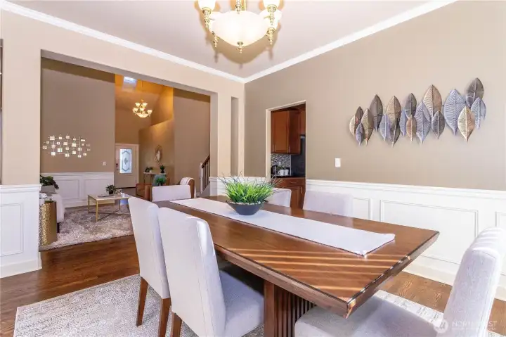 Elegant dining room with wainscoting and crown molding, open to the foyer and kitchen for effortless entertaining. A neutral palette and natural light highlight the rich wood floors, while metal leaf wall art adds a modern touch.