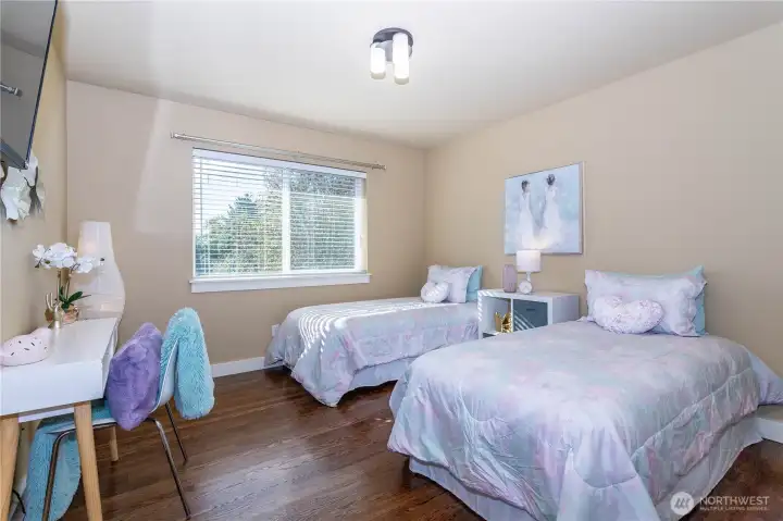 Bright and cheerful bedroom with two twin beds, perfect for kids or guests. The large window fills the room with natural light and looks out over the quiet neighborhood. Warm paint tones and hardwood floors create a calm, welcoming feel that blends perfectly with the home’s timeless design.