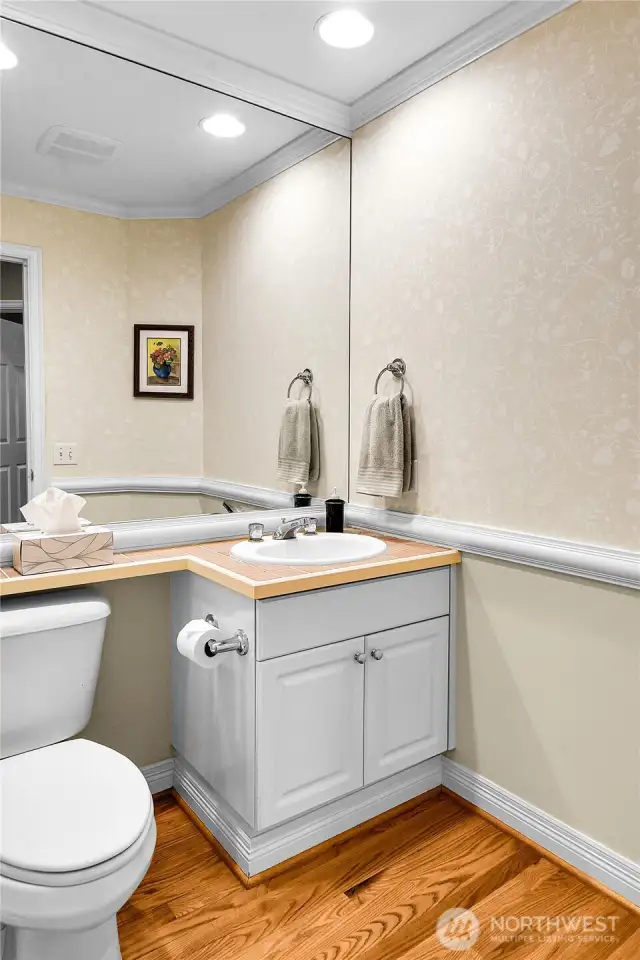 Powder bath with Oak-hardwood floor. Crown molding. Chair-rail wainscoting with elegant wallpaper above. Paneled-cabinet vanity with Banjo-style tiled counter, oval sink with brass and chrome fixtures. Full half wall vanity mirror. And elegant wallpaper above. Elongated watercloset.