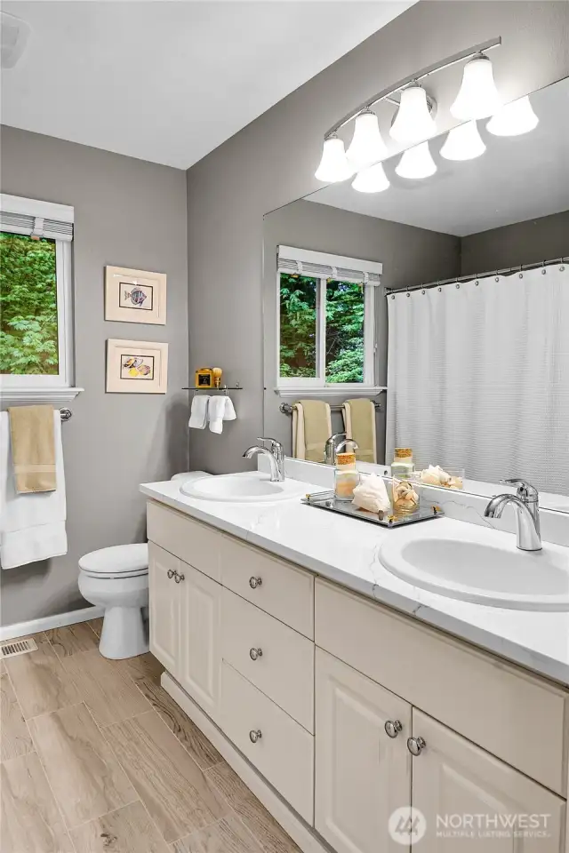 Main bath. Quartz counter with oval-shaped double sinks, chrome faucets. Mirror with bell shaped Alabaster style shades for vanity lights above. Window with screen two-inch blinds, looking out to the woods behind. Mansfield® standard watercloset. Bathtub with shower surround, privacy glass doors. Linen closet with tile floor, paneled Quaker door.