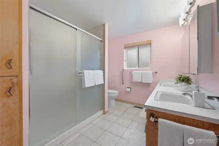 Main bathroom features a spacious walk-in shower with sliding glass doors and a wide vanity with dual sinks for everyday convenience. A frosted window provides natural light and privacy, while the generous layout offers comfortable room to move.