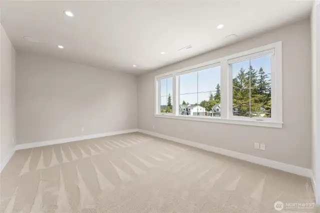 Spacious upstairs bedroom with plush carpet, recessed lighting, and a wall of windows offering treetop views and natural light.