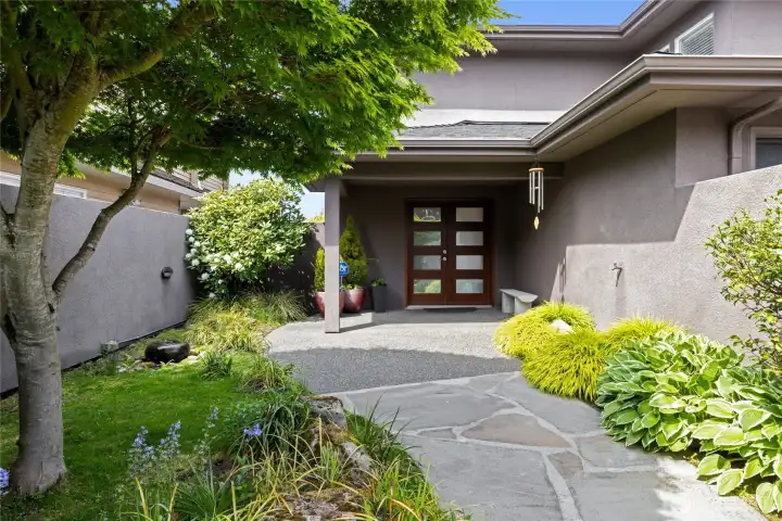 Inviting front porch with modern double-door entry, tranquil plantings, and thoughtfully designed pathways for a peaceful first impression. Welcoming covered entry framed by lush, mature landscaping and custom stonework, creating a serene, garden-like arrival experience.