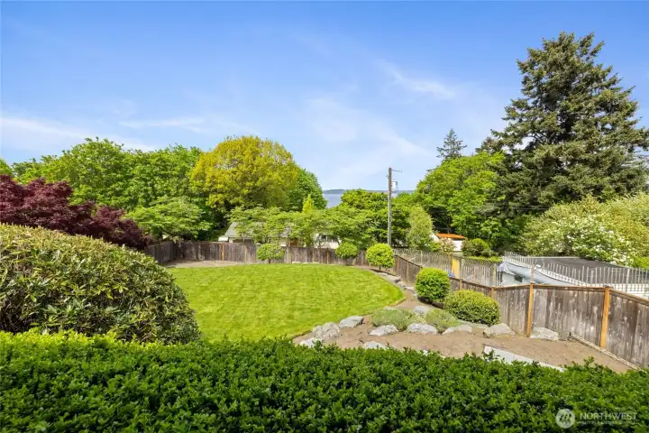 Beautifully landscaped backyard featuring terraced stonework, manicured lawns, and mature plantings creating a private, park-like setting. Expansive, level lawn framed by established greenery with peek-a-boo water views—perfect for play, relaxation, or future outdoor enhancements.