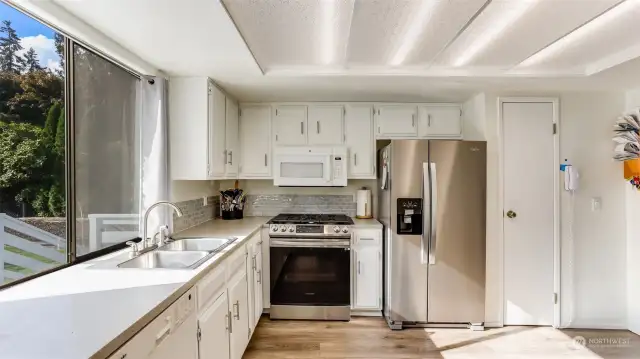 A spacious kitchen featuring white cabinetry, big window to the back yard, pantry space and room for small table.