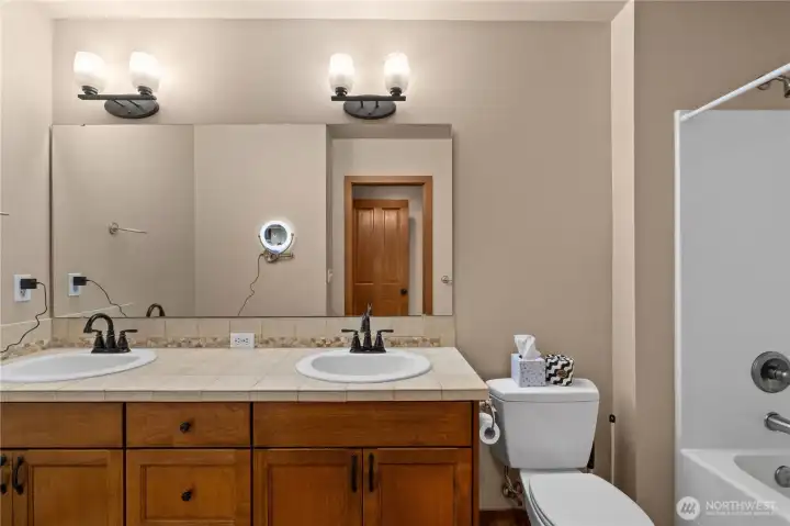 The en suite primary bathroom has a double vanity with ceramic tile countertop, a wall-mounted lit magnifying mirror and a deep bath/shower.