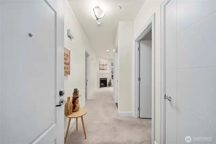 This hallway view highlights the smooth flow from the entry into the open living space, with soft carpeting and overhead lighting guiding you toward the fireplace and kitchen. The layout creates a seamless connection between the home’s functional and gathering areas.