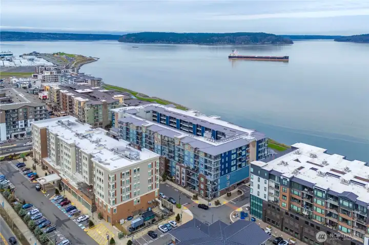A sweeping aerial view showcases the Baker Building along the shores of Commencement Bay, highlighting its exceptional location within the vibrant Point Ruston waterfront community. Enjoy immediate access to shoreline trails, seaside dining, and year-round outdoor recreation.