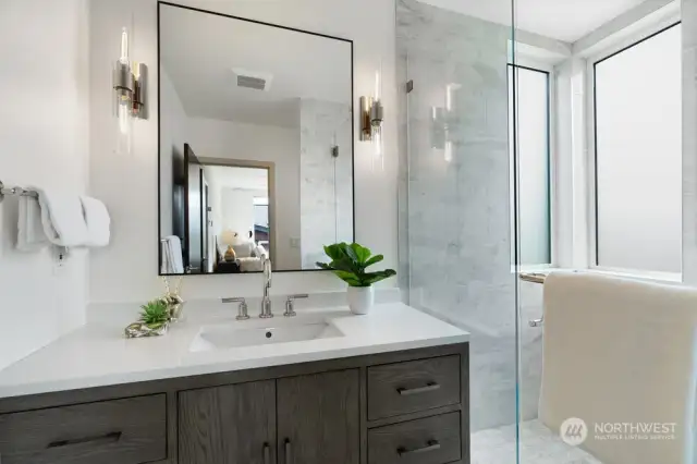 The primary suite bathroom has been updated with large 12x24-inch tiled floor, a large frameless glass tiled shower and furniture styled quartz vanity flanked with wall sconces.