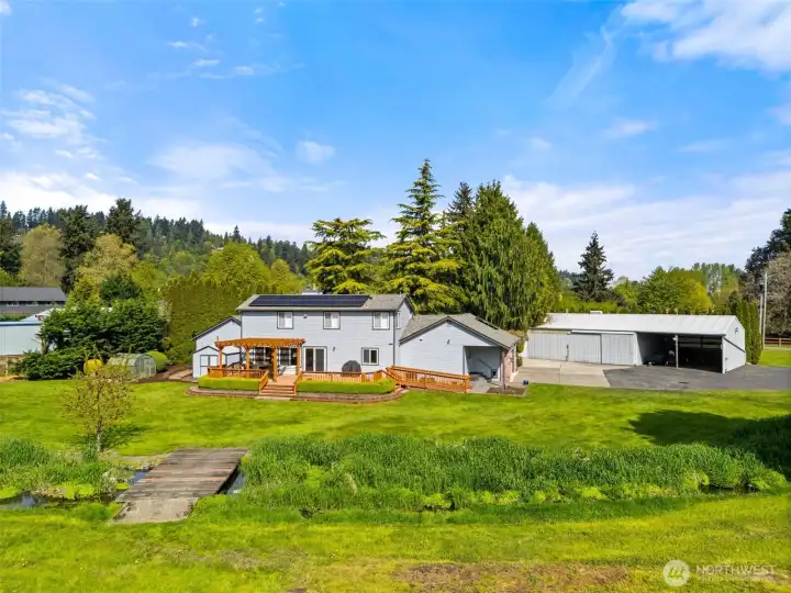 Aerial view of the back of the home showing the rear deck, detached garage/shop with 2 car carport, greenhouse, garden shed and creek.