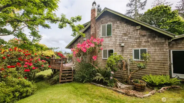 Framed by blooming rhododendrons and lush woodland textures, the exterior of this Nahcotta home is a perfect marriage of coastal charm and storybook whimsy. From the weathered shingles and cheerful chimney to the handcrafted garden path and driftwood details, every element welcomes you into a space where nature and comfort intertwine.