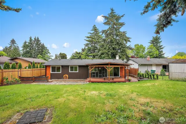 A full backyard view of the home showing off the spacious lawn, charming covered deck, and peaceful privacy framed by evergreens. Perfect for play, pets, or future garden beds!