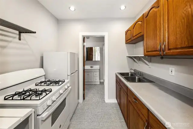 Apartment 1 Kitchen – Clean, Functional, Updated  Apartment 1’s galley kitchen balances utility and character, with newer flooring, updated lighting, and a classic gas range—ideal for serious cooks. Original wood cabinetry and thoughtful open shelving add warmth and storage, while a window at the far end brings in natural light. A well-equipped space within one of the building’s most architecturally significant apartments.