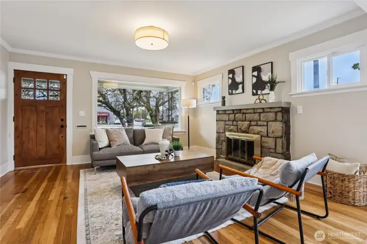 Light-filled living room with warm hardwoods and a statement fireplace creates an inviting space to gather. Large windows frame the outdoors, while the rich wood front door ties it all together with character and charm—comfortable, welcoming, and easy to fall in love with.