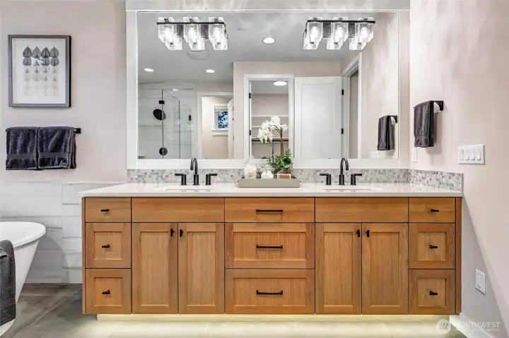 A beautifully finished primary bath featuring a wide double-sink vanity with warm wood cabinetry, quartz counters, and elegant mosaic backsplash detailing. Oversized framed mirror and sparkling contemporary lighting create a bright, spa-like atmosphere.