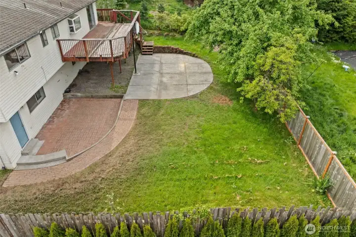 The aerial view reveals a substantial fenced lot with multiple distinct outdoor zones — brick paver patio, large curved concrete pad, elevated deck, and established trees along the perimeter.
