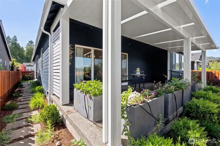 One of the things that sets this property apart is how beautifully the outdoor spaces have been planned. Oversized rectangular pavers create a clean, contemporary pathway along the side of the home, offering dry footing and elegant curb appeal at the same time. The combination of rich mulch beds, colorful plantings and evergreen accents brings texture and softness to the landscape without creating a maintenance burden. From this vantage point, you can appreciate how seamlessly the covered patio connects with the surrounding hardscape, creating an outdoor setting that feels both elevated and effortless.