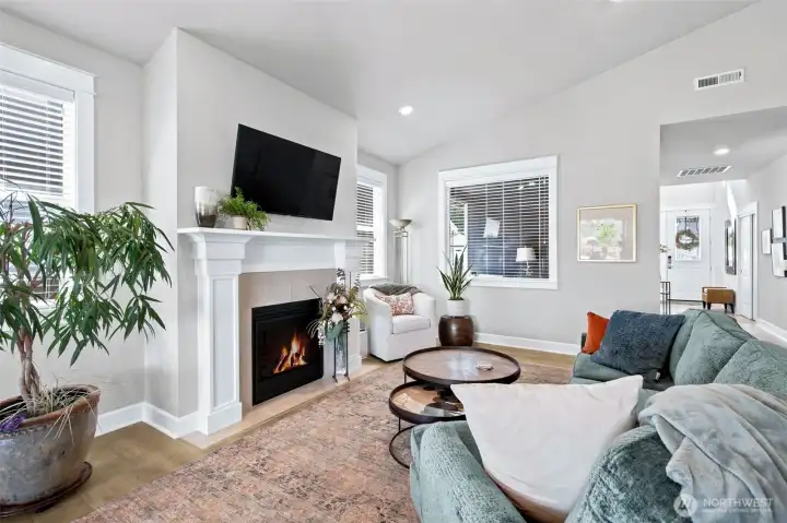 Anchoring the living room space is a gorgeous gas fireplace wrapped in a classic white mantel complete with elegant column details that feel straight out of a designer show home. Built-in media hookups above the mantel make entertaining easy, while matching windows on either side create symmetry and flood the room with natural light. The large window beyond offers a lovely view through the covered courtyard-style patio, adding depth and bringing the outdoors into the heart of the home. Between the soaring vaulted ceiling, warm flooring and beautifully balanced architecture, this room feels both grand and comfortable at the same time.