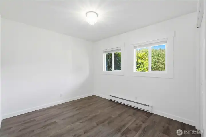 Bedroom with two windows facing mature trees in back yard, dark flooring, chandelier light, closet doors and baseboard heater. Bedroom with two windows facing mature trees in back yard, dark flooring, chandelier light, closet doors and baseboard heater.