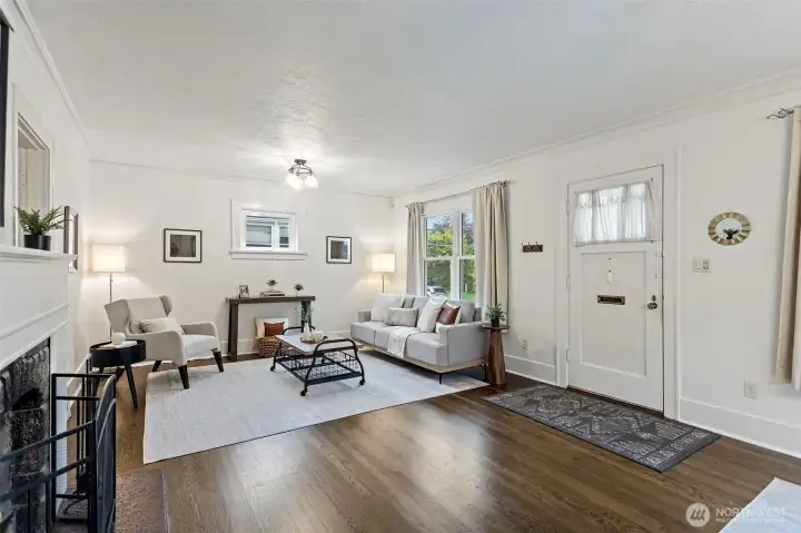 Bright and welcoming living room with newer hardwood flooring (2022), fresh paint, and updated lighting. Large windows bring in abundant natural light, complementing the charm of the cozy fireplace at the heart of the room.