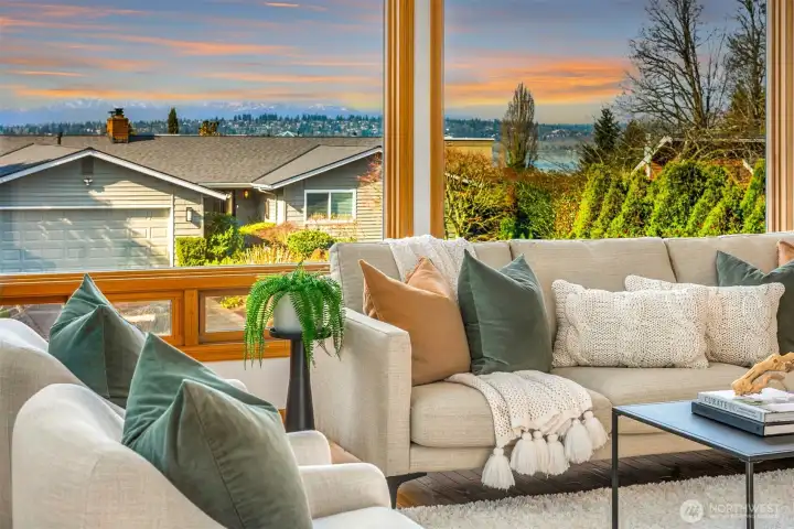 A sun-drenched living room with expansive windows framing panoramic views of lush greenery and the iconic Olympic Mountains beyond. Neutral tones, plush furnishings, and warm natural light create an inviting space where the indoors and the scenery seamlessly blend.