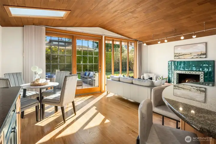 A seamless indoor-outdoor living space where the striking slatted wood ceiling, gleaming hardwood floors, and a cozy fireplace are bathed in natural light pouring through expansive glass doors — opening directly onto a lush deck and inviting the outdoors in at every turn.
