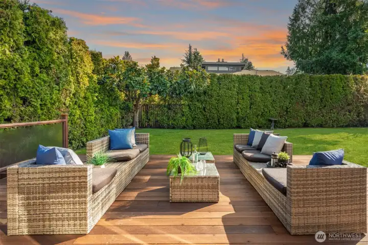 A beautifully private backyard oasis where the family room opens directly onto a generous deck with stylish outdoor seating, flowing seamlessly to a lush, flat lawn framed by mature trees and landscaping — a tranquil retreat perfectly suited for entertaining or quiet relaxation.