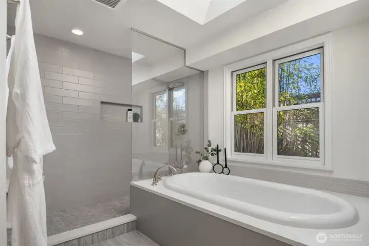 A beautifully updated primary bathroom showcasing a sleek walk-in shower with custom tile, a deep soaking tub framed by sun-filled windows looking out onto a serene, private bamboo garden — sophisticated grey and white finishes completing a spa-inspired retreat thoughtfully designed for everyday luxury.