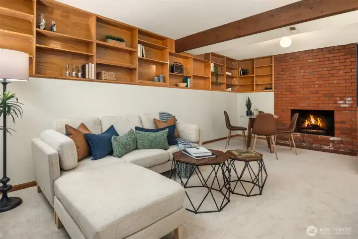 A spacious and inviting lower level rec room featuring a classic brick fireplace, warm wood built-in shelving, and plush comfortable seating — a versatile and welcoming space equally suited for movie nights, a playroom, or effortless conversion to additional living quarters.