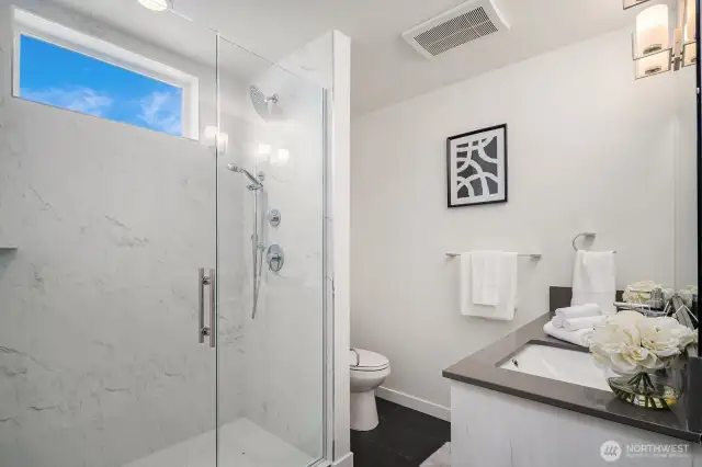 Experience spa-like opulence in this modern bathroom, featuring a glass-enclosed rain shower, dual vanities, and sleek cabinetry. An expansive window invites natural light, enhancing the tranquil atmosphere. (Photos are of staged model home.)