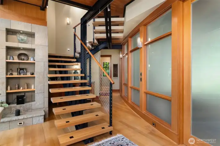 The simplicity of the floating staircase hovering above an enclave art wall. View to the curved wall of glass encapsulating the family room. The study is down the hall to the right and the primary suite is down the hall to the left.