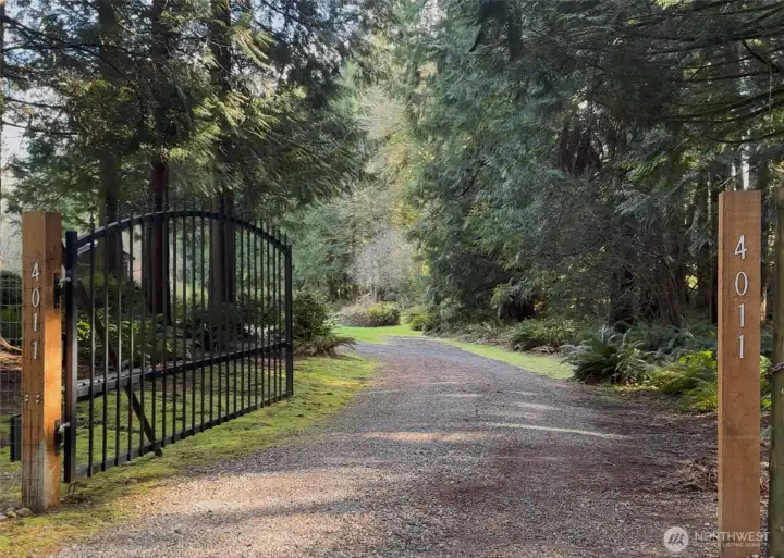 A gravel driveway stretches past the gate, winding gently through peaceful acreage beyond. The land feels expansive and secluded, offering privacy and tranquility.