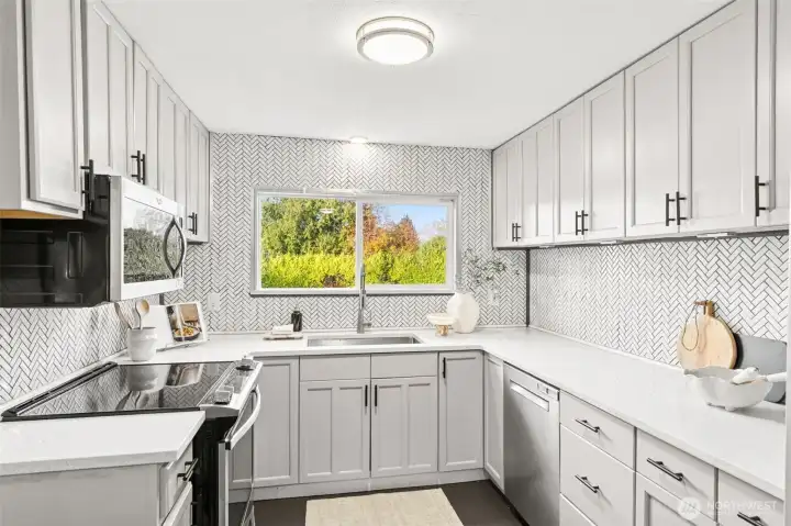 Stunning renovated kitchen at 4612 N 21st St, Tacoma, WA — showcasing quartz countertops, full herringbone tile backsplash, soft-close shaker cabinets, stainless steel appliances, and a large window with vibrant backyard views. This bright and spacious North Tacoma kitchen offers modern finishes, abundant storage, and a functional L-shaped layout perfect for cooking and entertaining.