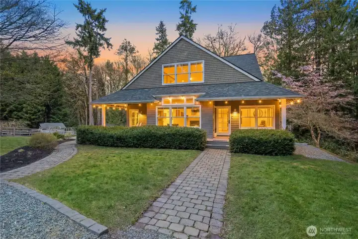 A classic Craftsman façade welcomes you with warm evening glow, crisp architectural lines, and an inviting covered front porch. Set against a backdrop of towering evergreens, the home sits privately on lush grounds with a paver walkway that hints at the peaceful, acreage lifestyle beyond.