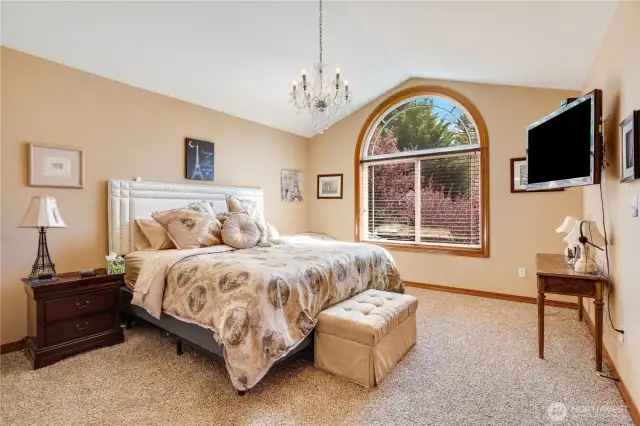 The vaulted ceilings elegantly highlight the spaciousness of this upstairs bedroom. The arched detail of the fully wood-wrapped window adds a sense of harmony to the room. Additionally, the walk-in closet offers organized storage solutions.