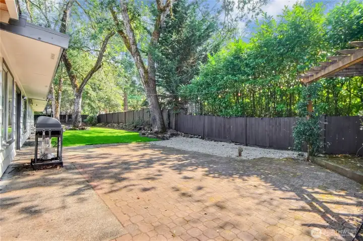 This inviting backyard features a spacious patio with string lighting, perfect for outdoor dining and entertaining. A pergola-covered seating area adds charm and shade, while the open paver space is ideal for barbecues or gatherings. Surrounded by lush greenery and a private fence, it offers both relaxation and privacy in a beautiful outdoor setting.