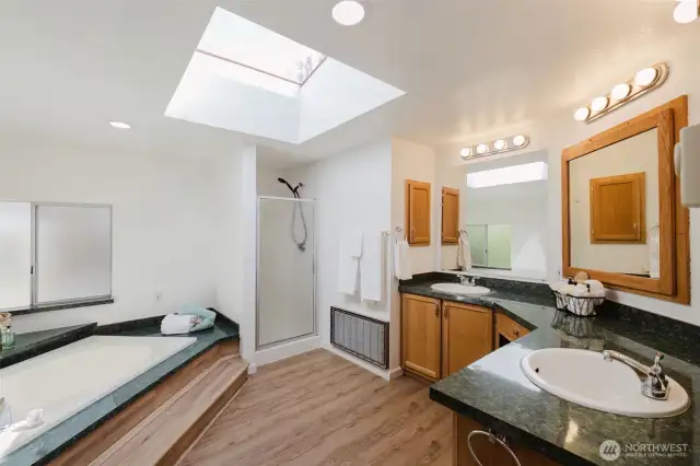 The generously sized primary bath features an L-shaped countertop with a sink at each end, providing ample counter space for each user. offering plenty of space for daily routines. Two steps lead to a relaxing soaking tub, complemented by a walk-in shower. Laminate wood floors complete the space, offering both style and functionality.
