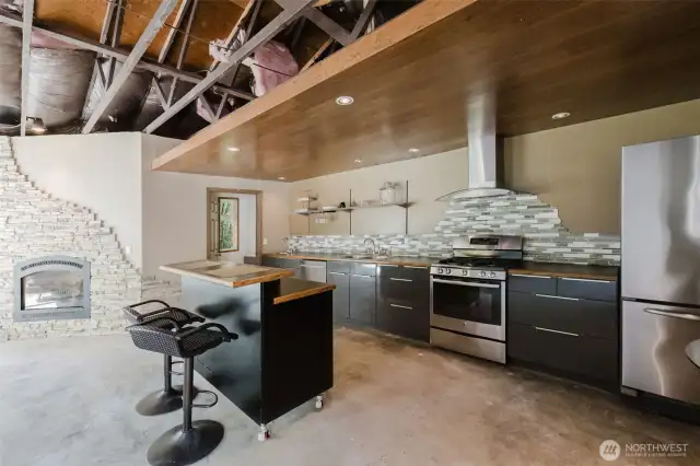 This guest studio perfectly blends modern comfort with a cozy, woodland vibe. A corner propane fireplace features a unique flat-rock surround that begins at the ceiling and flows down the wall in an organic, wavy pattern—adding both warmth and artistic flair. The kitchen is thoughtfully designed with black cabinets accented by stainless steel hardware, a tile backsplash, and stainless steel appliances. Overhead, a varnished wood ceiling with inset lighting adds warmth and character, completing the cabin-in-the-woods feel with a contemporary twist.