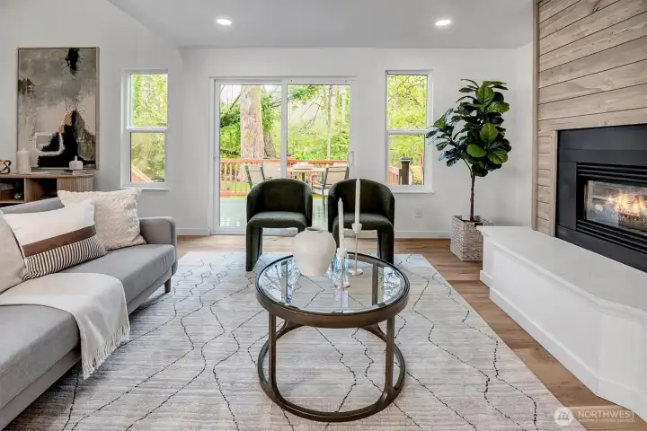 Living room featuring a wood-clad fireplace, laminate hardwood floors, and sliding glass doors opening to a deck with serene creek and tree-lined views.