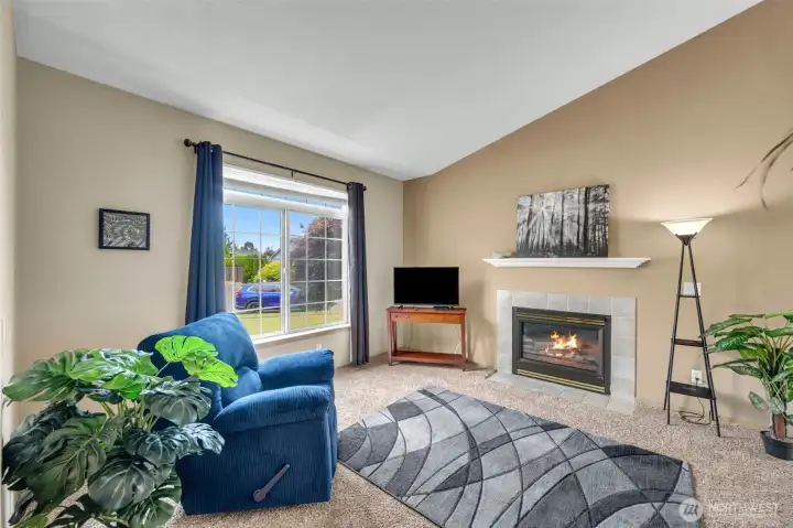 Bright and inviting living room featuring soaring vaulted ceilings, a cozy gas fireplace with tile surround and mantel, plush carpeting, and a large picture window that fills the space with natural light.