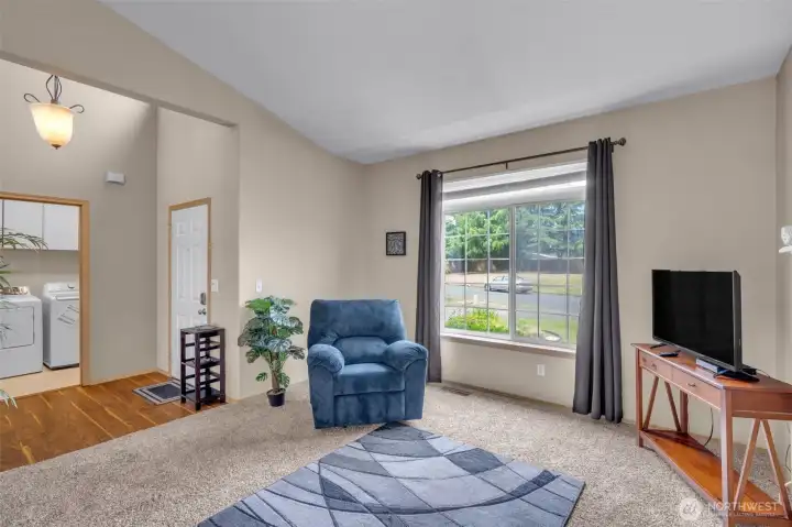 Welcoming living room with expansive front window, vaulted ceilings, and an open, comfortable layout. The front entry and adjacent laundry room offer convenient access while maintaining an inviting flow throughout the home.