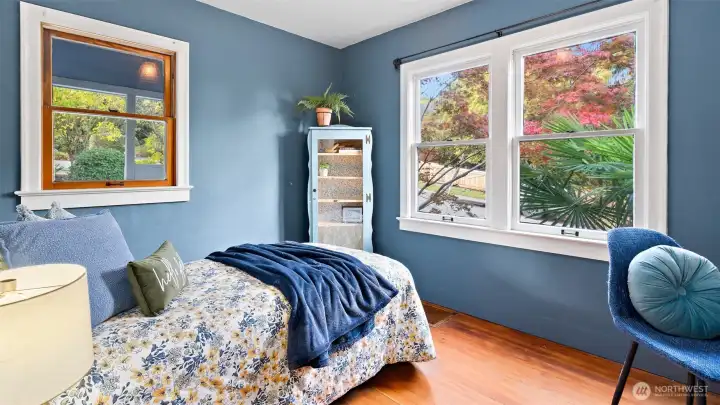This main-floor bedroom is full of charm and comfort, showcasing warm hardwood floors and windows on both sides that let in gentle natural light. It’s a bright, relaxing retreat that feels peaceful any time of day.
