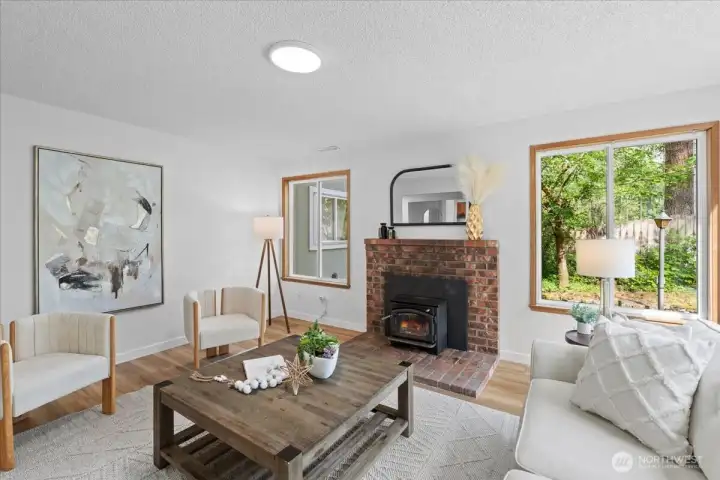 Living Room – Cozy gathering space centered around a brick fireplace with wood-burning stove insert. Large windows bring in natural light and views of the surrounding greenery.