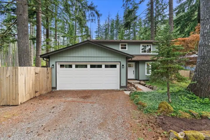 Front Exterior – Alternate view showcasing the home's attached garage, fenced side yard access, and surrounding greenery. Tall trees provide a picturesque backdrop throughout the property.
