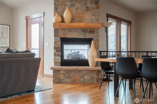 View of living and dining room w/ double sided stone fireplace, oversized windows and hardwood floors.
