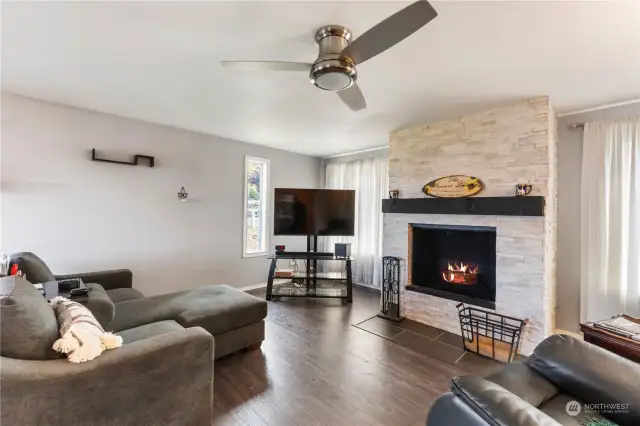 The great room features a fireplace with white brick surround, laminate wood flooring and ceiling fan.