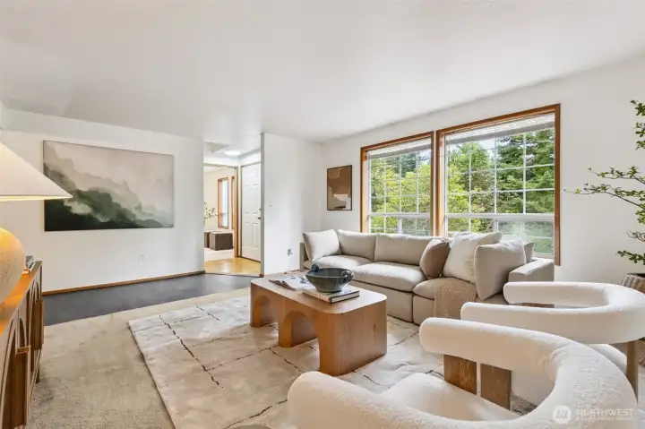 Sunlit living room with oversized windows framing lush forest views, featuring warm neutral décor and an inviting open layout that flows beautifully into the rest of the home.
