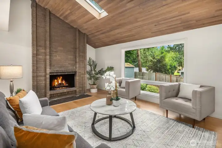 The living room's soaring vaulted ceiling, wood-planked skylight, and striking floor-to-ceiling fireplace combine to create a warm and architectural centerpiece that is truly one of a kind. Large windows frame the lush greenery outside, flooding this beautifully updated space with natural light and a seamless connection to the outdoors.