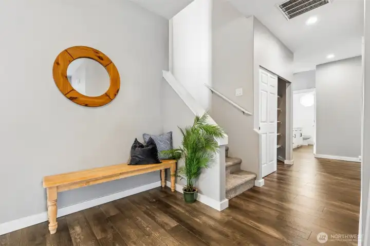 Wide plank hardwood floors in a rich, multi-tone grain pull you in immediately, paired with crisp white trim and a soft whisper-gray palette that keeps everything feeling fresh and elevated. The 9-foot ceilings hit right away, giving the space that open, expansive feel that just reads expensive. Stairs rise cleanly to the upper bonus level, while a full bath and coat storage sit thoughtfully in the background. It’s bright, composed, and sets the tone.